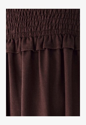 Brūns auduma tops ar savelkošu smokošanas detaļu, ruffled apakšmalu un mīkstu tekstūru, radot plūstošu dizainu ar smalkām krokām.