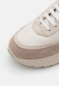 Beige och ljusbrun mockaskor med vita snören och gummisula, visade i närbild på en vit bakgrund.