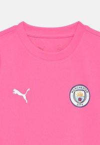 Ryškiai rožinė sportinė apranga su baltu Puma logotipu kairėje pusėje ir Mančesterio City futbolo klubo emblema dešinėje krūtinės pusėje.
