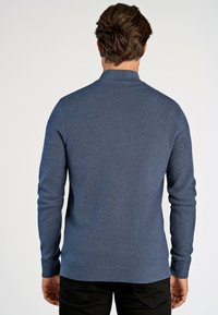 Niebieski sweter z dzianiny o strukturalnym wzorze i sztucznym kołnierzyku. Dopasowany krój z prążkowanymi mankietami i dołem. Czarne spodnie widoczne na dole.
