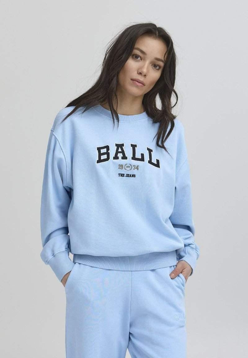 Lyseblå genser med avslappet passform, ribbestrikkede mansjetter og svart grafisk tekst "BALL 1974 THE JEANS" over fronten. Lomme detalj på buksene.