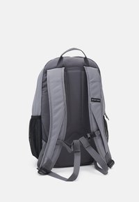 Gray fabric backpack with padded shoulder straps, mesh side pockets, and a hanging loop. Subtle stitching detail at back and logo patch.