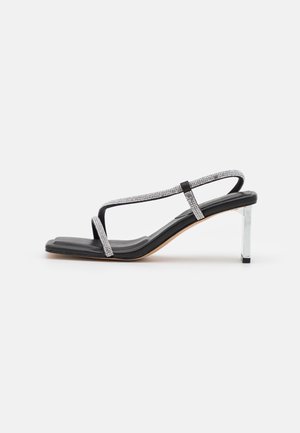 Sort sandal med tynde stropper pyntet med rhinsten, firkantet tå og en gennemsigtig høj hæl. Glat tekstur med en flad indersål.
