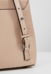 Coin d'un sac à main beige texturé avec une sangle ajustable, une boucle en argent et des détails bordés de noir sur un fond gris clair uni.