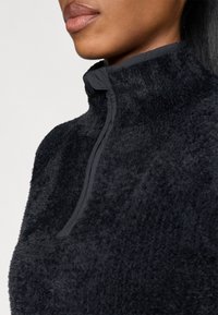 Sort fleece-pullover med høj krave og delvis lynlås. Stoffet er blødt, struktureret og ser varmt ud, med en glat kant langs halsudskæringen.