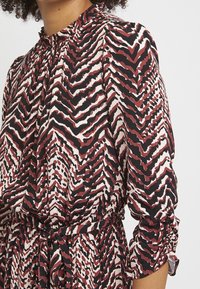 Röd och svart mönstrad klänning med hög halsringning och rynkade ärmar. Tillverkad av lättviktig tyg med ett strukturerat zigzag-mönster.