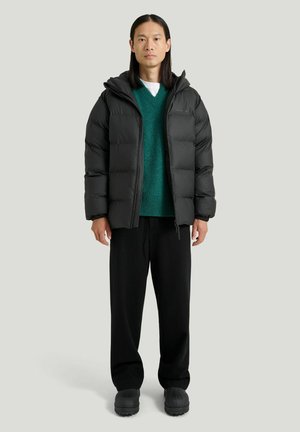 Voksen med langt hår iført en sort dunjakke over en grøn sweater, sorte bukser og sorte sko, stående mod en ensfarvet baggrund.
