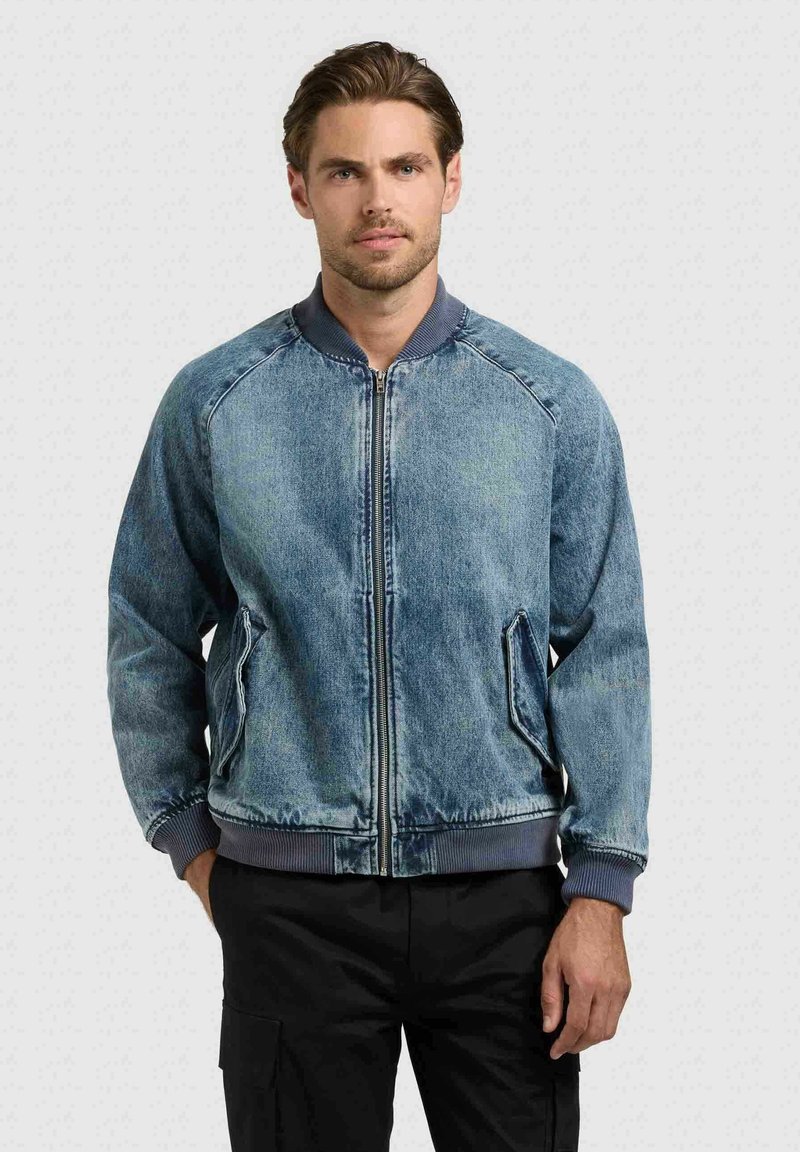 Homme aux cheveux courts brun portant une veste en jean bleue zippée et un pantalon noir, debout devant un fond clair uni.