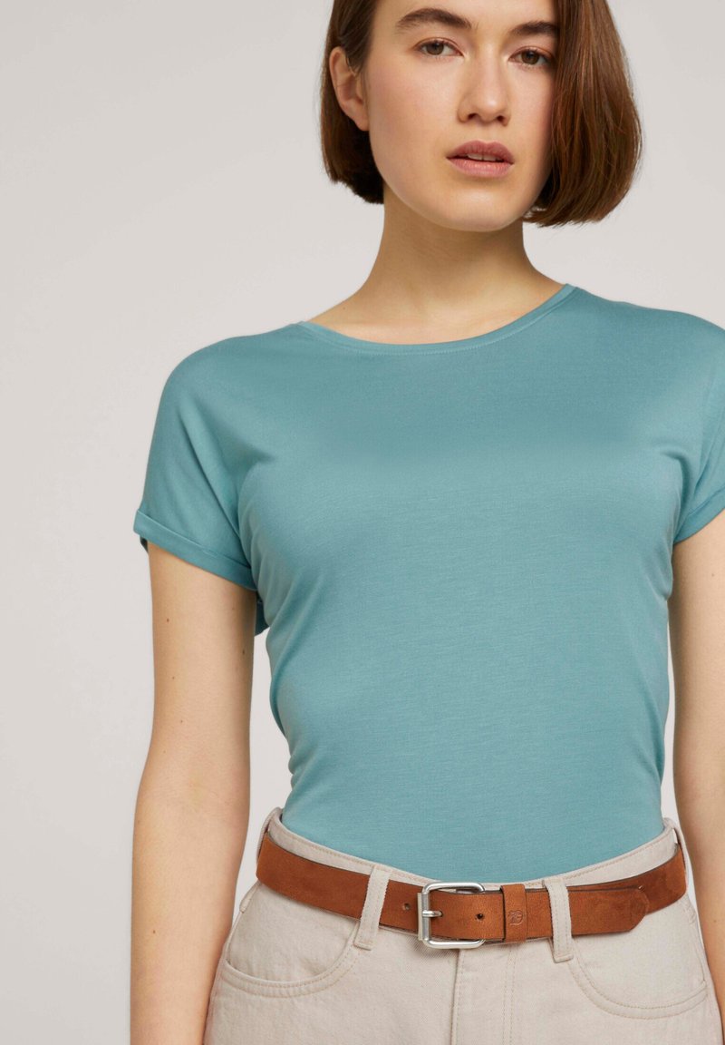 T-shirt bleu clair à manches courtes en tissu lisse, associé à un pantalon beige et une ceinture en cuir marron avec une boucle en argent.