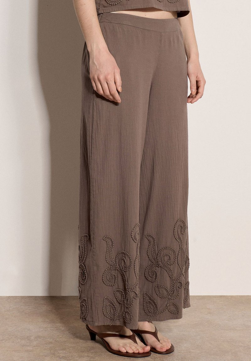 Femme portant un pantalon marron ample avec une broderie florale texturée à l’ourlet, associée à des sandales marron à talons, debout à l’intérieur.