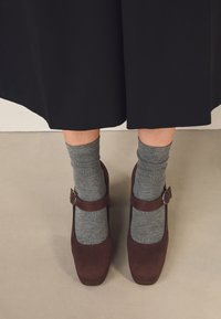 Bruna Mary Jane-skor i mocka med rund tå och justerbar rem. Bärs med gråa ankelsockor, mot en neutral golvbakgrund.