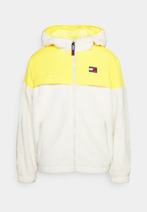 Person trägt eine gelb-weiße Fleece-Kapuzenjacke mit Reißverschluss, ein schwarzes Shirt, blaue Jeans und weiße Sneakers und steht vor einem schlichten weißen Hintergrund.