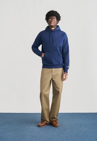 Μπλε-navy βαμβακερό φούτερ με κουκούλα με κορδόνι και μπροστινή τσέπη, συνδυασμένο με μπεζ παντελόνι και καφέ παπούτσια, στέκεται σε μπλε χαλί.