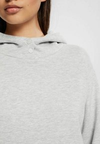 Felpa grigia chiara con una texture morbida, caratterizzata da una vestibilità ampia, un cappuccio con coulisse e due dettagli con bottoni a pressione sul colletto.