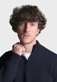 Námořnický pletený pulover s vysokým límcem a polovičním zipem vpředu, s žebrovanými manžetami a jemným logem na hrudi.