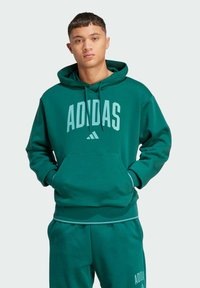 Haut de survêtement en coton vert avec poche avant, cordons de serrage et logo "ADIDAS" en bleu clair sur la poitrine. Joggers verts assortis visibles.