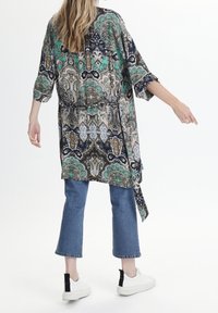 Kvinna stående med ryggen vänd, iförd en mönstrad lång tunika, korta blå jeans och vita sneakers med svarta hälflikar.