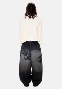 Άτομο με μακριά dreadlocks που φοράει άνετο λευκό πουκάμισο με μακριά μανίκια και φαρδιά μαύρα παντελόνια με διαβαθμισμένες υφές και σχέδια, στέκεται γυρισμένο προς τα πίσω.