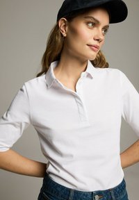 Biała koszulka polo z krótkim rękawem, z zapięciem na guziki i miękką strukturą, zestawiona z ciemnoniebieskimi dżinsami. Model nosi czapkę w kolorze czarnym.