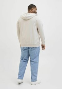Svijetlosmeđi zip-up hoodie s mekanom teksturom i kapuljačom, uparen s širokim plavim trapericama i bijelim tenisicama. Vidljivi detalji na leđima uključuju šavove.