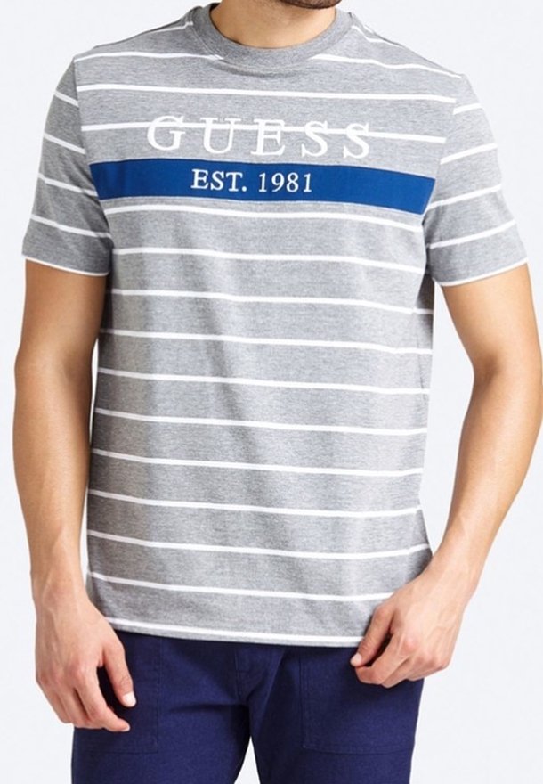 Homme portant un t-shirt à manches courtes rayé gris et blanc avec une bande bleue et le texte "GUESS EST. 1981", associé à un pantalon bleu foncé.