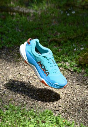 Lys blå løpesko med teksturert overdel, hvite og oransje såle accenter, som har en fremtredende "HOKA"-logo på siden og en hæltap.