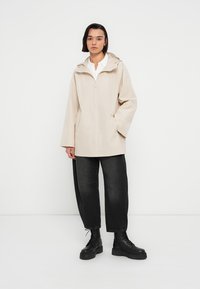 Veste imperméable beige à capuche, avec une fermeture éclair à l'avant, des poches latérales et une coupe ample, associée à un pantalon large noir et des bottes noires.