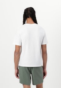 White cotton t-shirt with short sleeves and a round collar, paired with green shorts, viewed from the back. Plain design, no visible patterns.