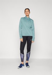 Lichtblauw groene lange mouwen rits top, structuurstof. Zwarte leggings met een blauw en paars abstract patroon en witte accenten. Sport schoenen.