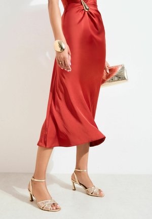 Woman wearing a red satin midi dress with gold high-heeled sandals, a large gold bracelet, and holding a gold clutch bag.