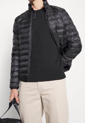 Mann trägt schwarze Steppjacke über schwarzem Pullover und beigen Hosen, hält schwarze Tasche in linker Hand vor weißem Hintergrund.