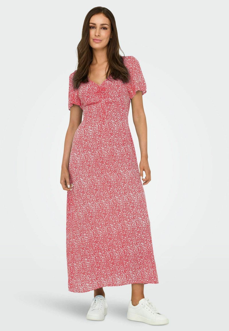 Abito maxi rosso a fiori con maniche corte a sbuffo, vita cintrata e silhouette fluida. Abbinato a scarpe da ginnastica bianche per un look casual.
