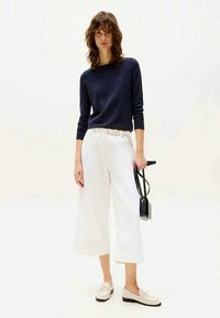 Marine crew-neck trui, witte wijde broek, beige loafers. Model draagt een kleine zwarte handtas. Eenvoudige, casual outfit.