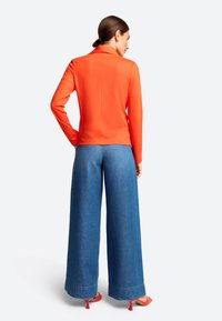 Leuchtend orangefarbenes Langarmoberteil mit einem dezenten Kragen, kombiniert mit weit geschnittenen blauen Jeanshosen und roten Absatzsandalen. Glatte Textur und taillierter Schnitt.