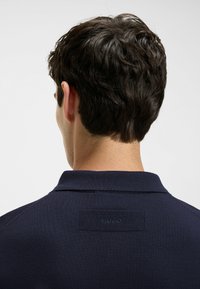 Mężczyzna z krótkimi ciemnymi włosami, ubrany w granatową koszulę z kołnierzykiem, z teksturowanym prostokątnym naszywką z napisem "HUGO" na górnej części pleców.