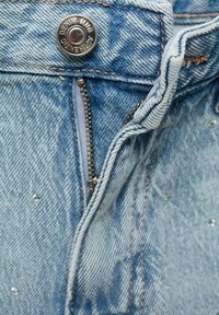 Svetlo modra jeans tkanina z sprednjim zadrgo, srebrnim gumbom in subtilnimi okraski v obliki majhnih sijočih žebljičkov.