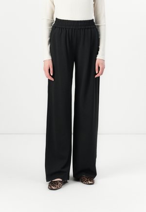 Personne portant un pantalon large noir, un haut blanc à manches longues côtelé et des chaussures plates à imprimé léopard, debout devant un fond blanc uni.