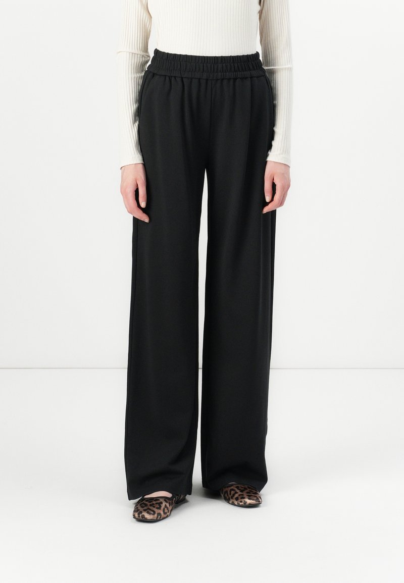 Personne portant un pantalon large noir, un haut blanc à manches longues côtelé et des chaussures plates à imprimé léopard, debout devant un fond blanc uni.