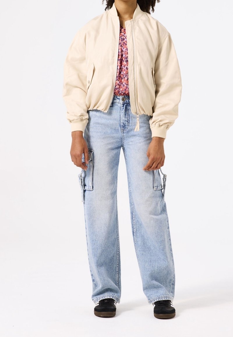 Beżowa kurtka typu bomber na kwiecistym topie, do tego jasnoniebieskie jeansy cargo z dużymi kieszeniami i czarne sneakersy. Prosty design, luźny krój.