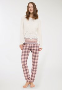 Giovane donna in top bianco a maniche lunghe con una grafica di un gatto che dorme e libri, e pantaloni del pigiama a quadretti rosa e grigi, a piedi nudi.