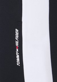 Navy ja valkoiset puuvillasekoitekankaiset housut, joissa on pystysuora valkoinen raita ja painettu "TOMMY HILFIGER" -logo valkoisella punaisilla korostuksilla.