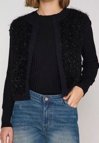 Mujer con un cárdigan sin mangas negro y peludo sobre una camiseta de manga larga negra de canalé y jeans azules desgastados, con la mano en el bolsillo.