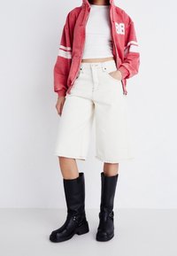 Sudadera roja con cremallera y rayas blancas, top corto blanco, pantalones cortos Bermuda crema y botas negras altas con hebillas.