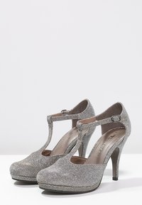 Scarpe con tacco alto argento con platform, design a T, texture glitterata e punta rotonda. Chiusura con fibbia alla caviglia.