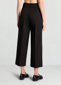 Pantalon large noir à taille haute, en tissu lisse et aux lignes épurées. Une paire de chaussures noires visible au niveau de la cheville. Pas de poches apparentes.