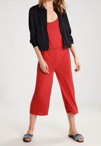 Mono rojo con piernas cortas, cintura elástica, diseño en capas, combinado con una chaqueta bomber negra y sandalias de tejido azul.