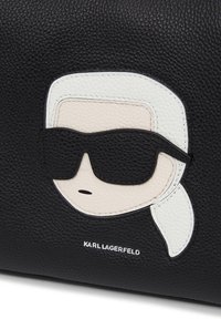 Sort tekstureret lædertaske med et grafisk ansigt i hvid og beige nuancer, solbriller og teksten "KARL LAGERFELD" nederst.