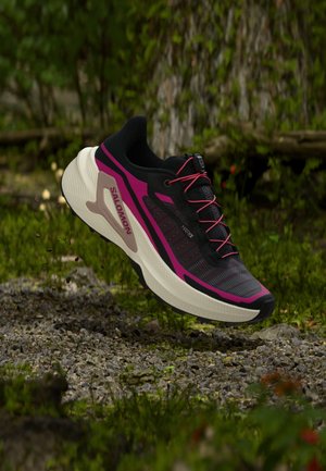 Chaussure de course Salomon noire et violette avec des lacets roses flottant au-dessus d'un sol forestier gravillonneux et mousseux.