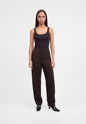 Femme debout, face à l’avant, portant un haut noir sans manches, un pantalon marron foncé taille haute et des chaussures à talons noires sur fond blanc.
