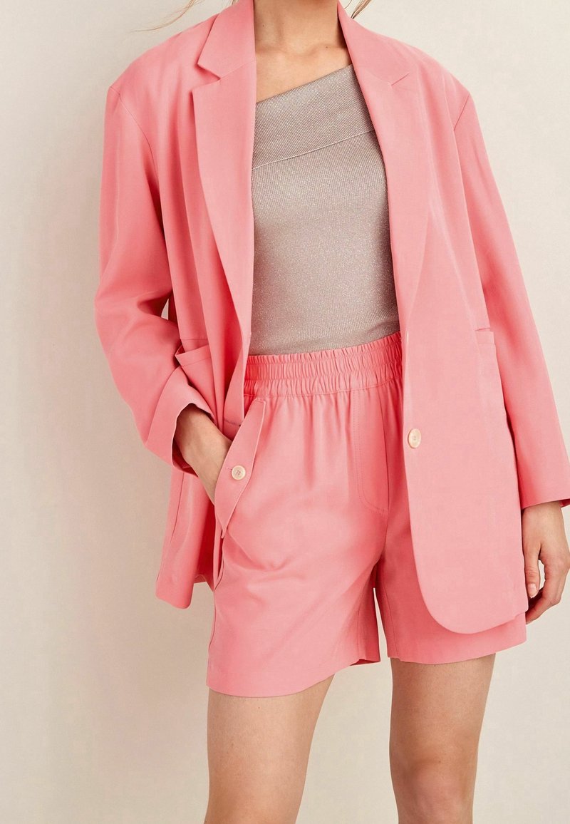 Blazer rosa de corte relajado, combinado con pantalones cortos rosa a juego. El blazer cuenta con solapas muesca y acentos de botón. Parte superior texturada gris por debajo.